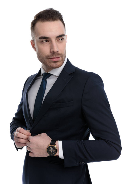 Charming businessman adjusting jacket and looking over shoulder while standing on white studio background - Zdjęcie, obraz