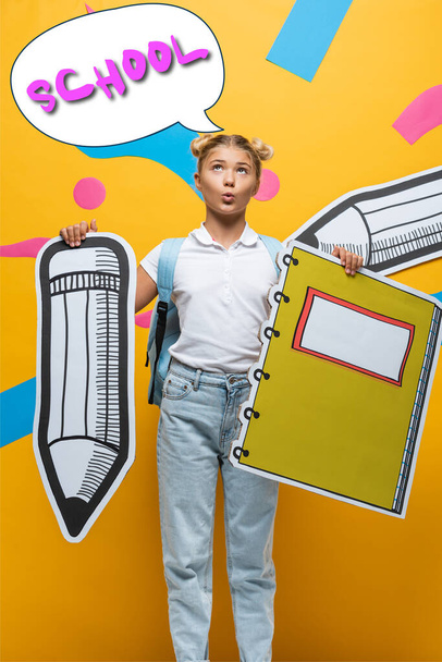 Pensive schoolgirl holding paper pencil and notebook near speech bubble illustration with school lettering on yellow  - Foto, Imagen