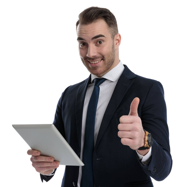Positive businessman giving thumbs up and holding tablet, smiling while standing on white studio background - Valokuva, kuva