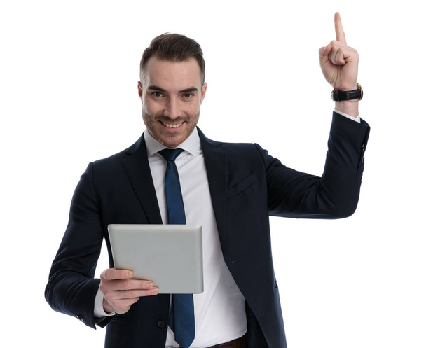 Ευτυχισμένος επιχειρηματίας κρατώντας tablet και δείχνοντας προς τα πάνω, χαμογελώντας ενώ στέκεται σε λευκό φόντο στούντιο - Φωτογραφία, εικόνα