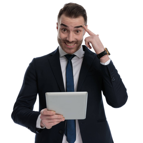 Веселый бизнесмен, держащий планшет и указывающий на лоб, стоя на белом фоне студии - Фото, изображение