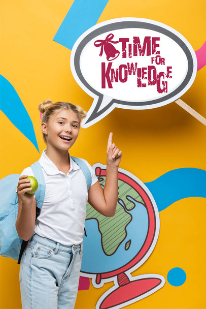 Школьник с рюкзаком и яблоком указывает на бумажный пузырь речи со временем для написания знаний на желтом  - Фото, изображение