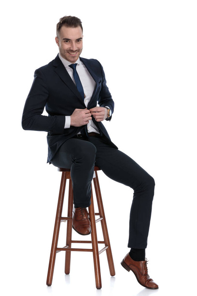 Positivo uomo d'affari che ride e regola la giacca mentre siede su uno sgabello su sfondo bianco da studio - Foto, immagini