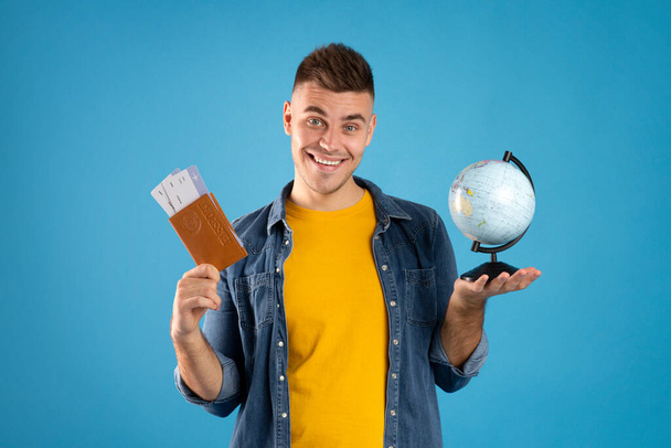 Усміхнений чоловік мандрівник тримає глобус, паспорт і квитки на літак на синьому фоні студії
 - Фото, зображення