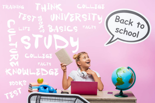Schoolgirl holding book and looking at speech bubble with back to school lettering near gadgets, globe and illustration on pink  - Fotó, kép