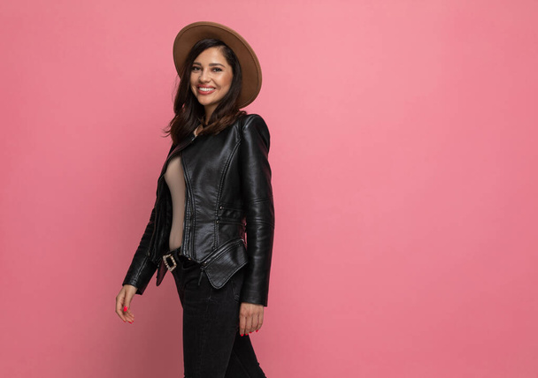 Side view of happy fashion model smiling, wearing hat while standing on pink studio background - Photo, Image