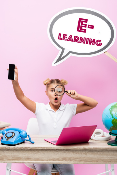 schoolgirl holding magnifying glass and smartphone with blank screen near speech bubble with e-learning lettering, globe and laptop on pink  - Foto, Imagen