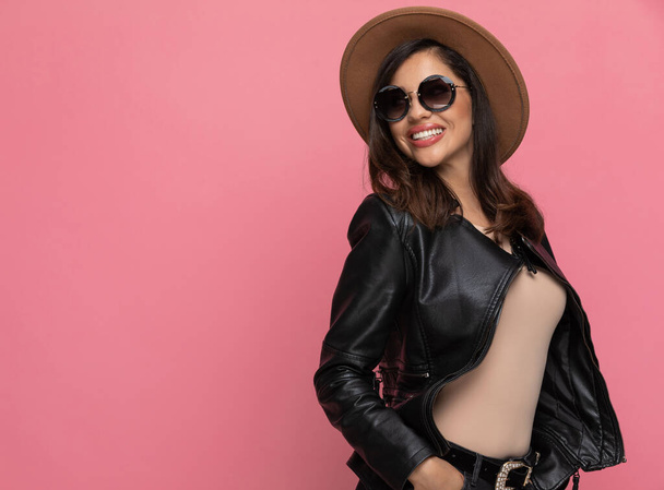 Позитивная модель, смеющаяся руками в карманах, в шляпе и солнцезащитных очках, стоя на розовом фоне студии - Фото, изображение