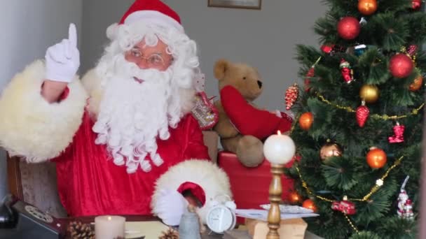 adult santa claus in red suit sits at table, Christmas tree is beautifully decorated with balls, garlands, concept of christmas, new year celebration, holiday sales and discounts - Video, Çekim