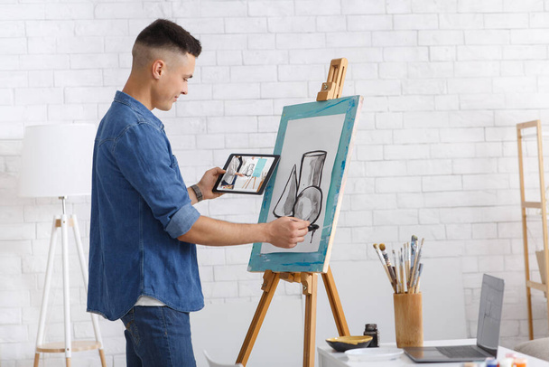 Art lesson online and hobbies during coronavirus quarantine at home. Young man draws picture, holds tablet in hand and watches training video - 写真・画像
