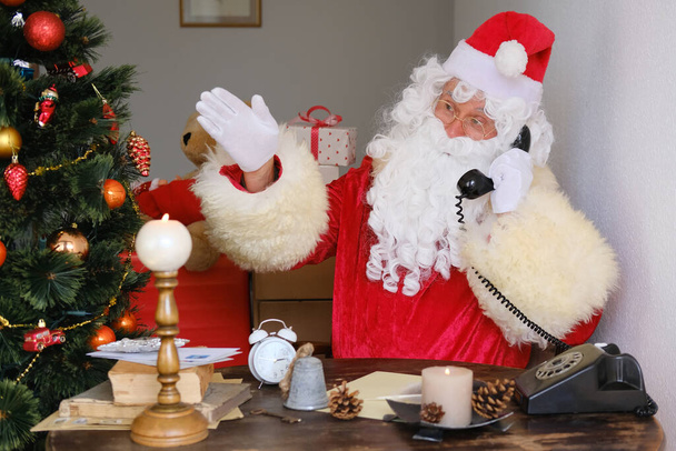 adult santa claus in red suit sits at the table, calls on old phone, tree is beautifully decorated with balls, the concept of christmas, new year celebration, holiday sales and discounts, sylvester - Фото, изображение