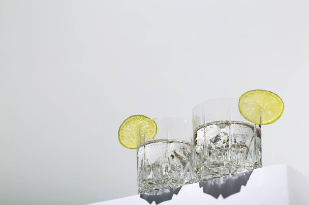 Vodka with ice and lime in two glasses on white stand. Hero view, shadows and sunlight trends. Copy space - Fotó, kép