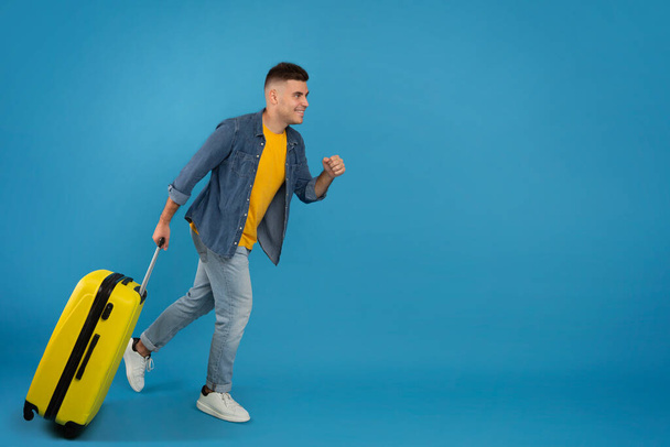 Ganzes Porträt eines fröhlichen männlichen Touristen mit Gepäck, das auf den Kopierraum vor blauem Studiohintergrund zuläuft - Foto, Bild