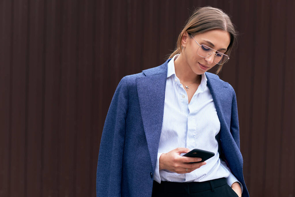 Female entrepreneur in elegant blue suit and optical eyewear texting email letter on mobile phone using Internet. Businesswoman messaging for discussing time of meeting using modern mobile phone - Foto, imagen