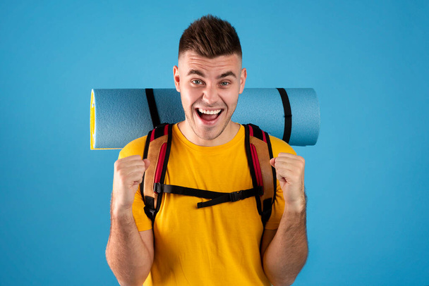 Excited young man with tourist equipment making YES gesture over blue studio background - Photo, Image