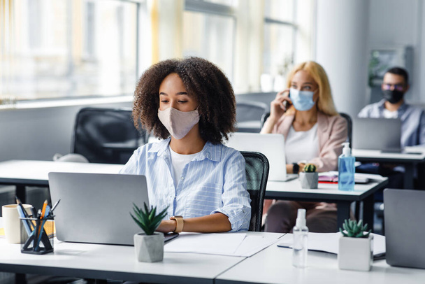 Social distancing at coronavirus outbreak situation. Pretty african american lady in protective mask typing on laptop, working for company support center - Photo, Image