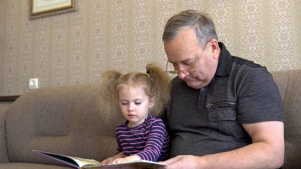 Kleindochter leest een boek met opa. Het meisje fronst naar het boek en luistert aandachtig naar grootvader. Zittend op de bank samen close-up - Foto, afbeelding