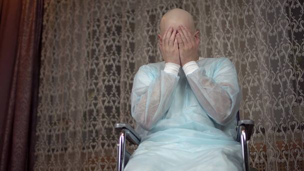 Лисий молодий чоловік з раком дивиться на камеру і плаче. Пацієнт прикриває обличчя руками, сидячи в інвалідному візку вдома. Випадання волосся завдяки хіміотерапії
. - Фото, зображення