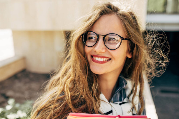 Closeup portrait of a smiling young student woman wearing transparent eyeglasses standing next to the college campus and carrying lots of books and folders on a sunny day. - Foto, afbeelding