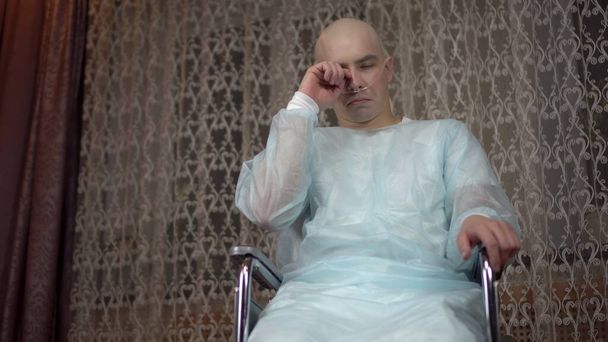 Egy kopasz fiatalember az onkológiával a kamerába néz. A beteg letörli a könnyeket, miközben otthon ül egy tolószékben. Kemoterápia miatti hajhullás. - Fotó, kép