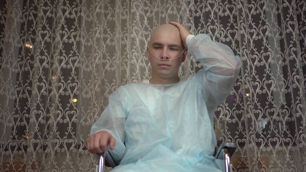 Een kale jongeman, een patiënt met oncologie, kijkt jammer genoeg in de camera. Een patiënt raakt een kaal hoofd aan terwijl hij thuis in een rolstoel zit. Haaruitval als gevolg van chemotherapie. - Foto, afbeelding