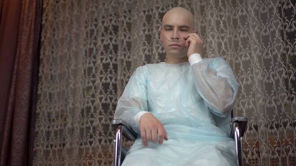 Onkolojisi olan kel genç bir adam kameraya üzgün bir şekilde bakar. Hasta evde tekerlekli sandalyede otururken solunum cihazını ayarlar. Kemoterapi yüzünden saç kaybı.. - Fotoğraf, Görsel