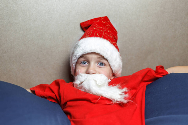 A blue-eyed boy with a Santa hat and an artificial white beard sitting on a bag chair. Portrait on a brown background - Photo, Image