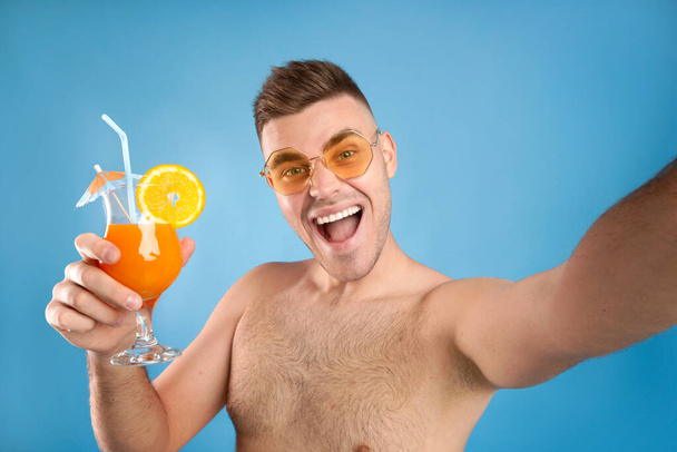 Opgewonden jonge man in een zonnebril houden verfrissende zomer cocktail en het nemen van selfie op blauwe studio achtergrond - Foto, afbeelding