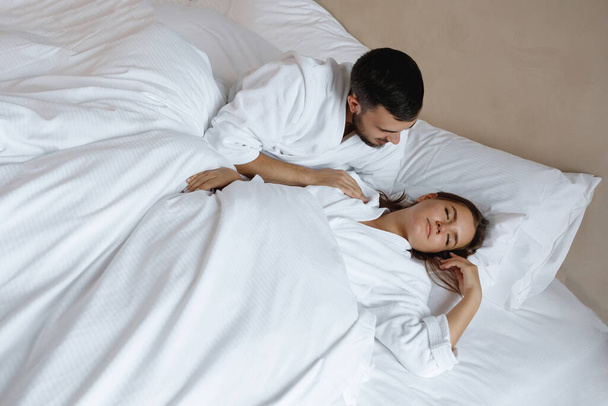 ベッドでカップル。若いひげを生やした男と女は白い枕の上に横になり、早朝に目を覚ます. - 写真・画像
