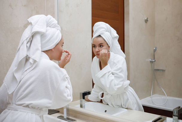 Молодая женщина в белом халате стирается в раковине с краном в ванной комнате. Отражение в зеркале. Выбранный фокус. - Фото, изображение