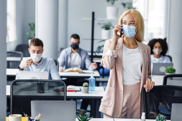 Coronavirüs salgını sırasında yeni normal ve sağlıklı koruma. Koruyucu maskeli bir iş kadını ofis içi iş arkadaşlarıyla telefonda konuşuyor. - Fotoğraf, Görsel