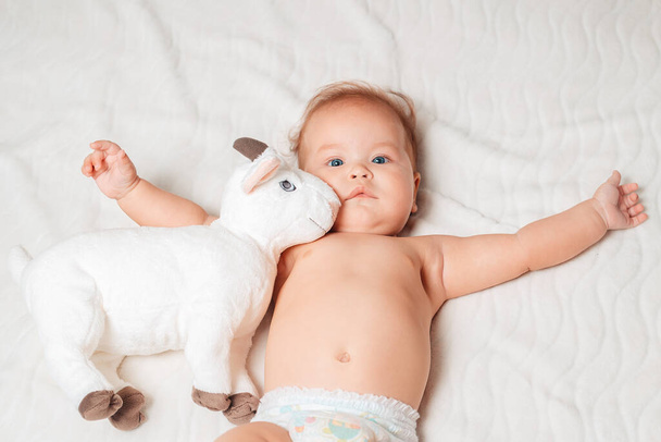 Retrato de un lindo bebé en pañales acostado en una sábana blanca con un juguete de peluche, brazos extendidos en diferentes direcciones. Vista desde arriba. Concepto de una infancia feliz. - Foto, imagen