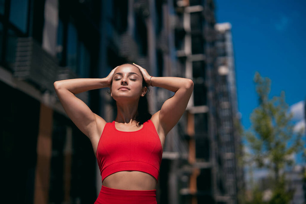 Fit young woman with dark ponytails wearing red sportswear. Sporty girl with arms raised touching her hair and smiling. Blurred city background. Apartment block and trees in background. - Φωτογραφία, εικόνα