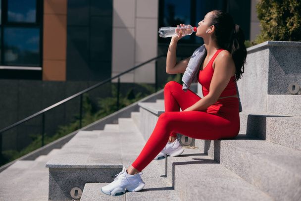 Side view of atletic young woman wearing red top and leggins nd white sneakers sitting on stairs. Girl taking break and drinking water from plastic bottle. Urban sport life concept. - Foto, Imagen