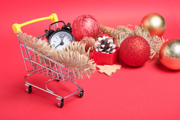 christmas decor and black small alarm clock in a mini shopping trolley on a red background, new year shopping concept - Fotoğraf, Görsel