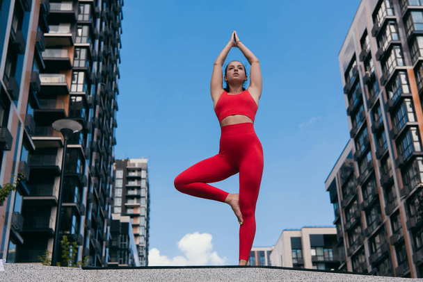 Mooie goed gebouwde vrouw die in Tree Yoga Pose staat. Meisje draagt rode sport top en leggins evenwichtig in yoga positie met opgeheven handen. Laag hoekschot. Stedelijk sportconcept. - Foto, afbeelding