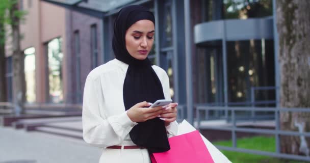 Arabic young woman in traditional headscarves looking at phone walking at street. Pretty muslim female in hijabs reading messages, scrolling bad news on smartphone after shopping. - Кадры, видео