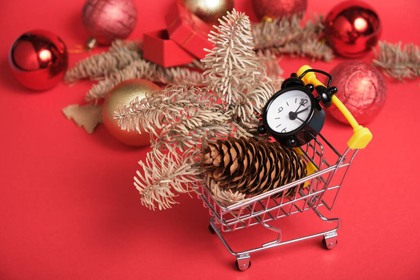 christmas decor and black small alarm clock in a mini shopping trolley on a red background, new year shopping concept - Fotoğraf, Görsel