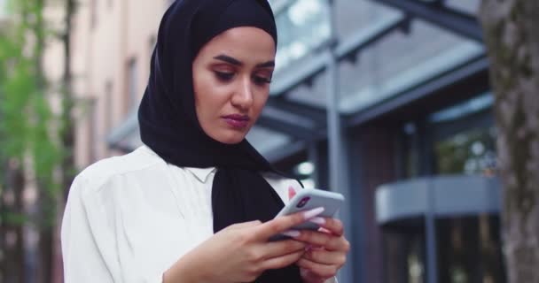 Close up retrato de mulher árabe em lenços de cabeça tradicionais olhando para o telefone e sorrindo. Mulher muito muçulmana em hijabs ler mensagens, mensagens de texto, navegar na Internet no smartphone depois de fazer compras. - Filmagem, Vídeo