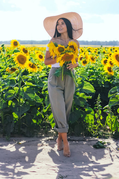 A beautiful brunette woman in a white top on a field with sunflowers holds flowers of sunflowers in her hands. Yellow sunflowers. - Foto, afbeelding