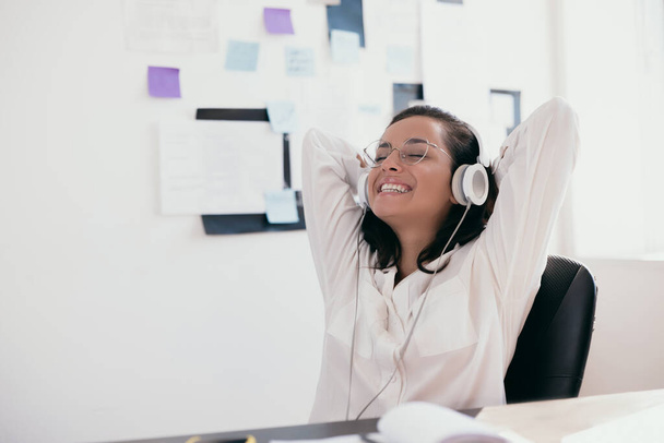Intelligent young woman with arms on head relaxes listening to music. Happy female office or freelance worker in white shirt with bright smile on blur background. - Фото, изображение