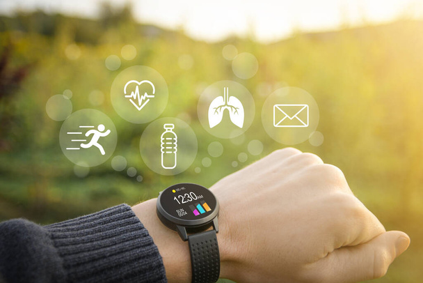Smart horloge, fitness tracker bij de hand in de buitenlucht op een wazig groene achtergrond met pictogrammen van basisfuncties.Concept van de technologie om de gezondheid te controleren. - Foto, afbeelding