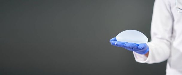 Panoramico di una protesi mammaria in silicone nelle mani di un medico. Dottore in mantello bianco che mostra una protesi mammaria su sfondo scuro. C'è ampio spazio per inserire testi. - Foto, immagini