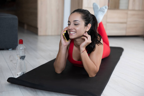 Mujer joven en chándal rojo haciendo ejercicio o yoga en casa. Chica bonita que se relaja después del entrenamiento o entrenamiento. Hablar inalámbrico en línea usando el teléfono inteligente. Tecnologías y dispositivos modernos. Después del entrenamiento. - Foto, imagen