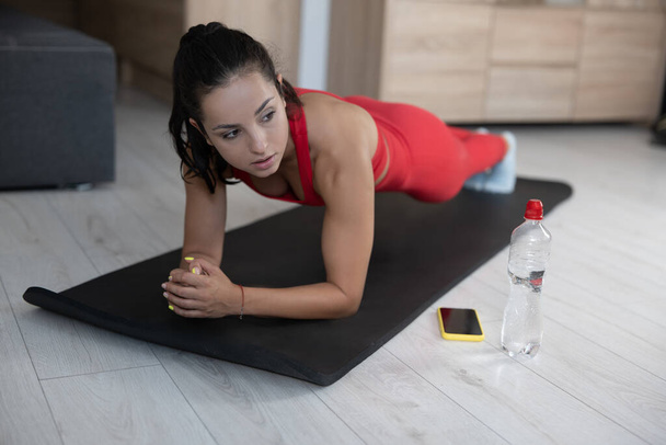 Young woman in red tracksuit doing exercise or yoga at home. Serious concentrated well-built girl standing alone in plank position on yoga mat. Looking to right. - Photo, Image