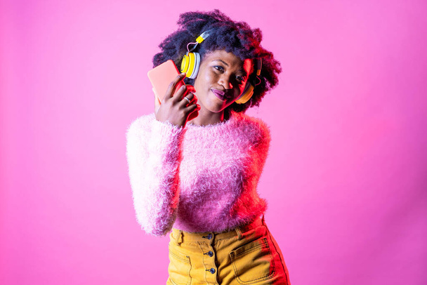 Young beautiful black woman dancing indoor on pink baground holding smartphone - Isolated diverse female clubbing listening streaming music - happiness, excitement, dancing concept - 写真・画像