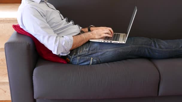 Мужчина в джинсах и рубашке использует ноутбук, лежащий на диване, когда ему звонят по мобильному телефону - Кадры, видео