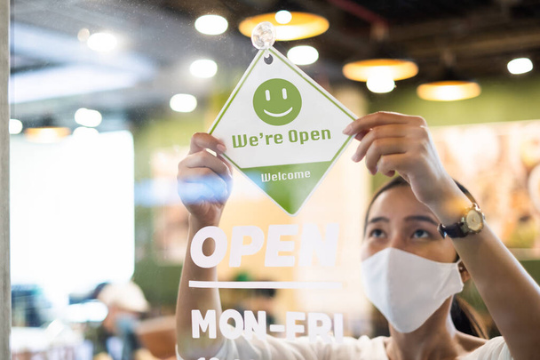 Propietaria de negocios mujer asiática usar máscara protectora ppe colgando letrero abierto en su restaurante / cafetería abierta de nuevo después de bloquear debido al brote de coronavirus covid-19 - Foto, Imagen