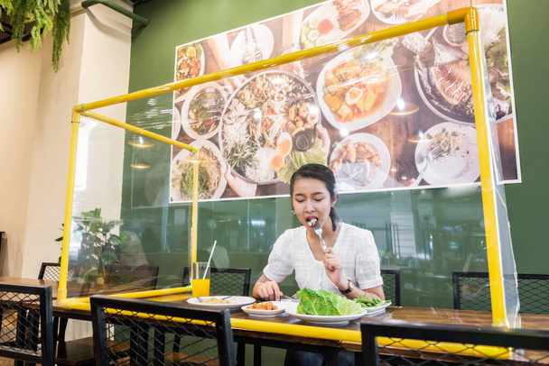 Asian woman sitting separated in restaurant eating food with table shield plastic partition to protect infection from coronavirus covid-19, restaurant and social distancing concept - Photo, Image