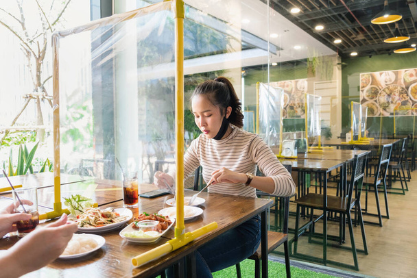 Mujer asiática sentada separada en restaurante comiendo comida con escudo de mesa partición plástica para proteger la infección del coronavirus covid-19, restaurante y concepto de distanciamiento social - Foto, Imagen